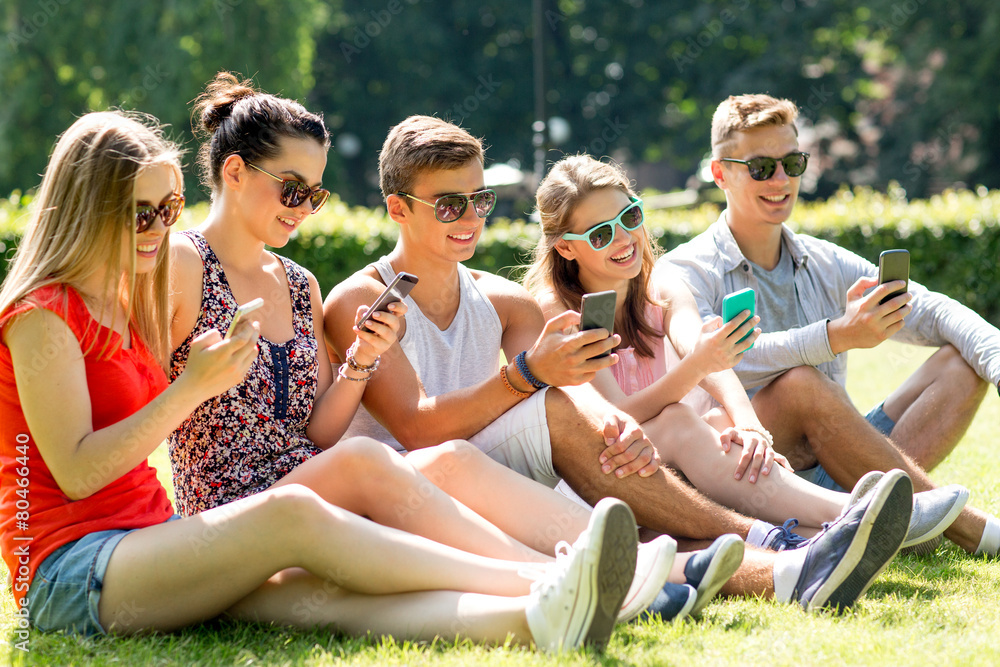 smiling friends with smartphones sitting on grass smiling friends with smartphones sitting on grass
