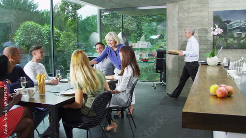 Happy group of family & friends sit down to eat a meal together in modern home