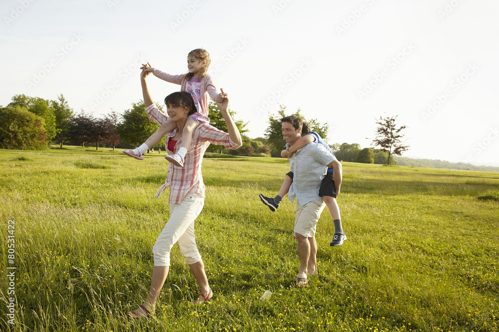 A family, two parents outdoors in the summer giving piggybacks to two children. A family, two parents outdoors in the summer giving piggybacks to two children.