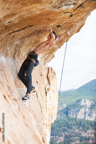 Young man clipping rope while clinging to cliff under ledge