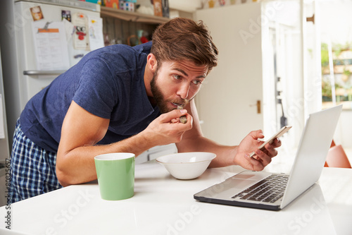 Man Eating Breakfast Whilst Using Mobile Phone And Laptop