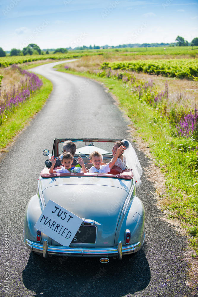 A newlywed couple is driving a retro car with their kids