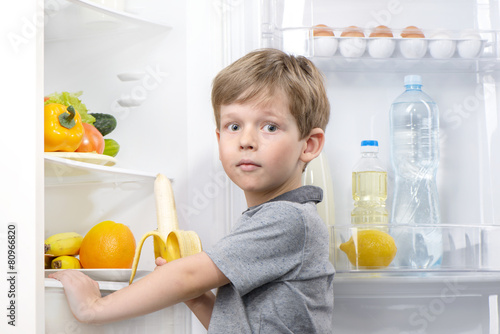 Little cute boy holding banana near open fridge