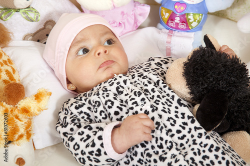 Bebita vestida con una pijama de animal print recostada en su cuna rodeada de animales de peluche photo