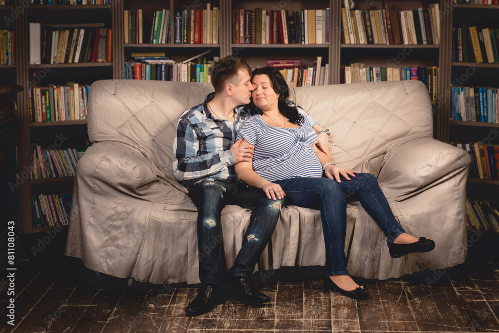 young pregnant couple hugging on sofa against bookshelves young pregnant couple hugging on sofa against bookshelves