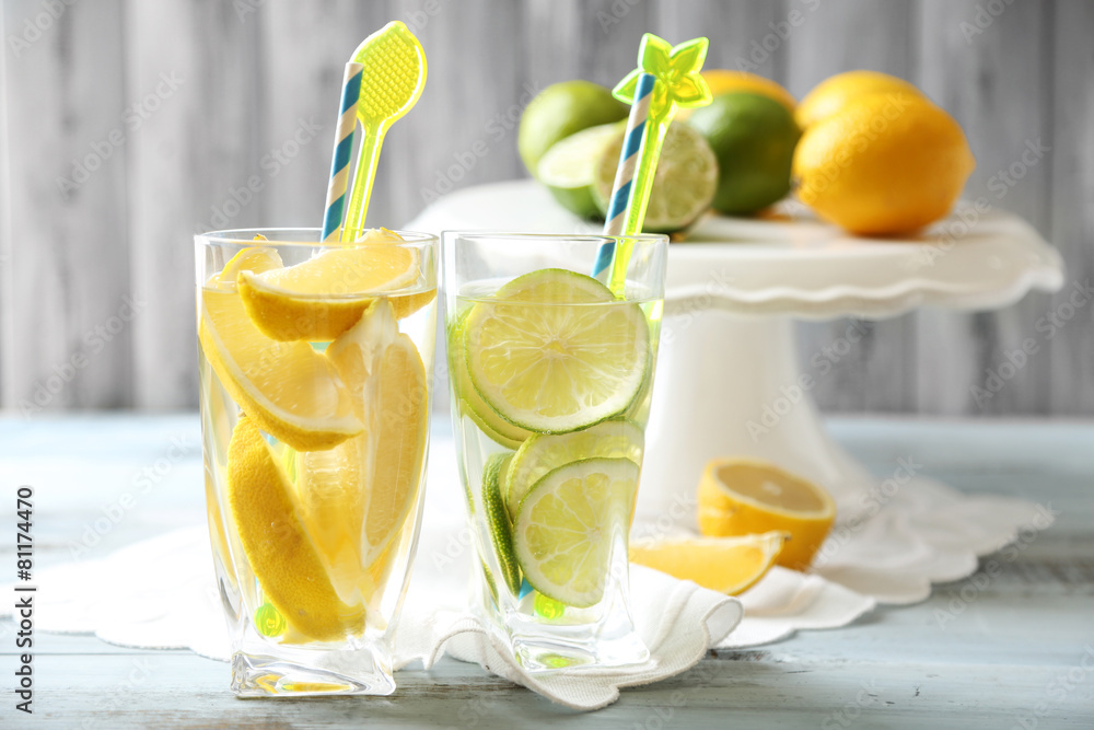 Fototapeta premium Refreshing lemon and lime infused water in glasses with citrus slices and decorative stirrers on a wooden table, the drinks are placed on a white napkin on a light wooden table