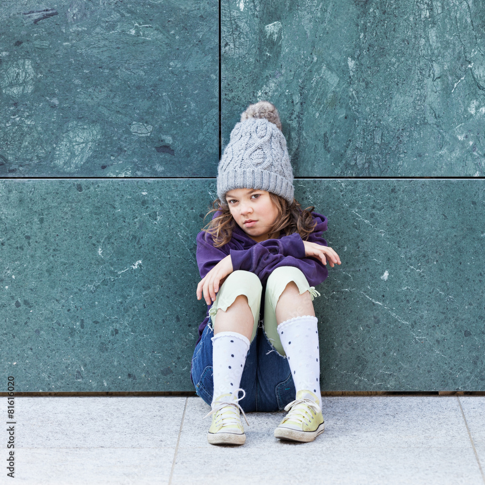 Portrait of a little girl with wool hat. Little girl sitting on the ground angry or not happy Portrait of a little girl with wool hat. Little girl sitting on the ground angry or not happy