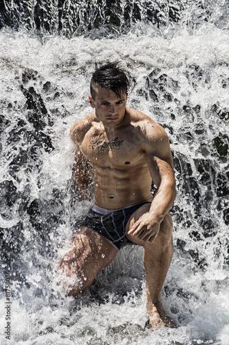 Attractive muscular shirtless young man under small waterfall