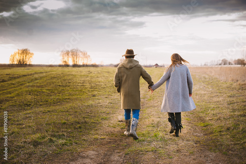 Beautiful couple in fashionable coat comes in a field at sunset,