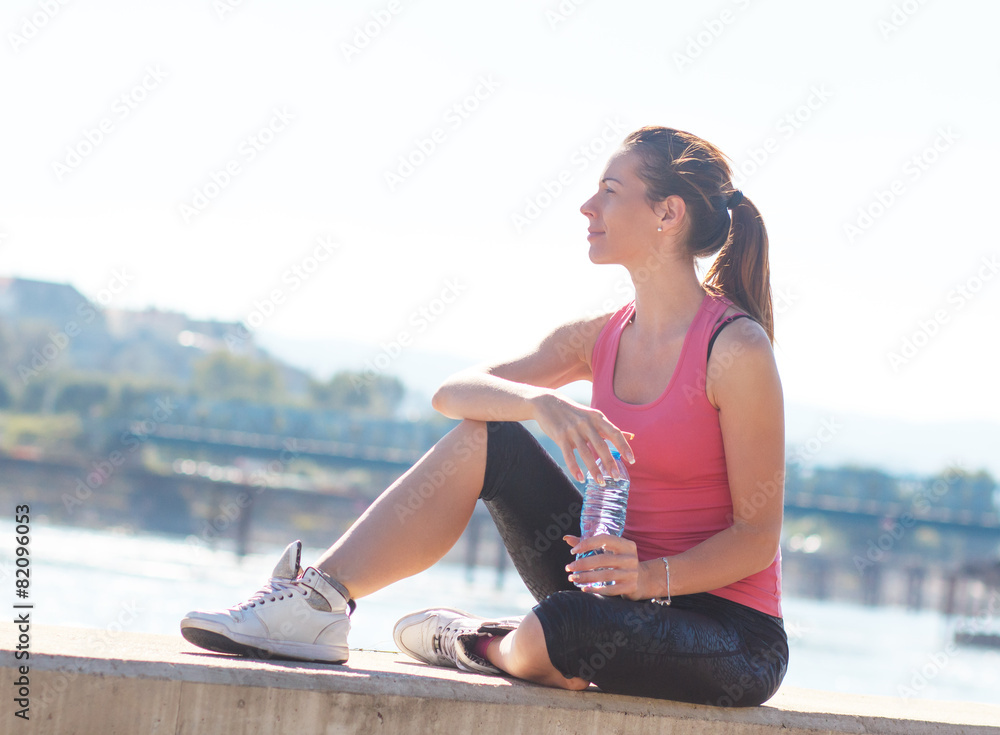 Fototapeta premium Young woman sitting on the riverbank and resting after jogging.