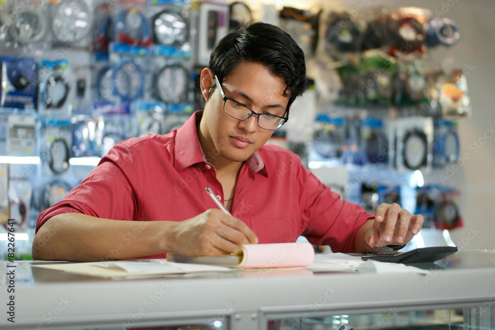 Chinese Man Working In Computer Shop Checking Bills And Taxes Chinese Man Working In Computer Shop Checking Bills And Taxes