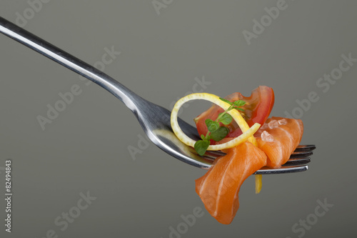 Salmon piece with lemon, tomatoe and herb arranged on fork.