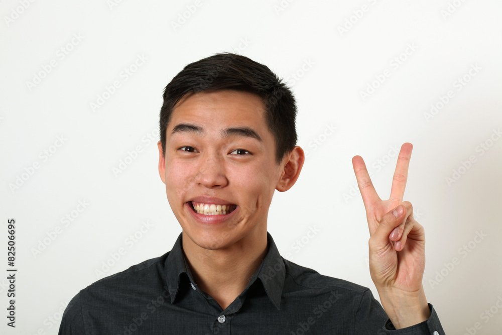 Smiling young Asian man giving the victory sign and looking at c Smiling young Asian man giving the victory sign and looking at c