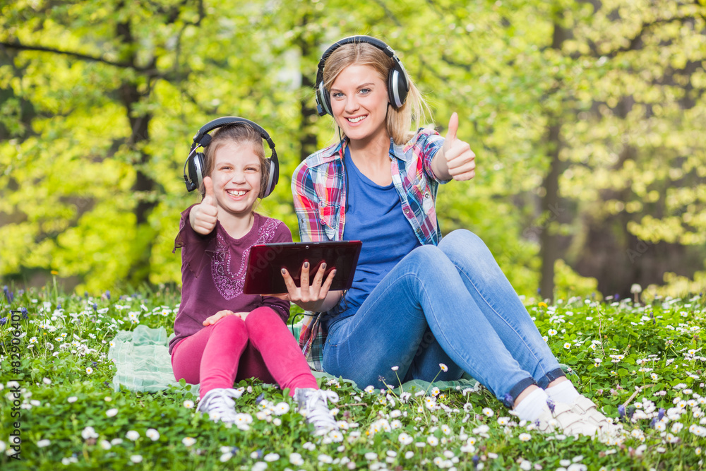 Fototapeta premium Sisters in park, using digital tablet and listening music