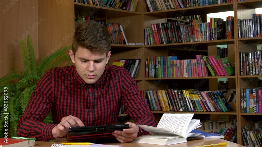 Student using his digital tablet in a library, preparing for Student using his digital tablet in a library, preparing for