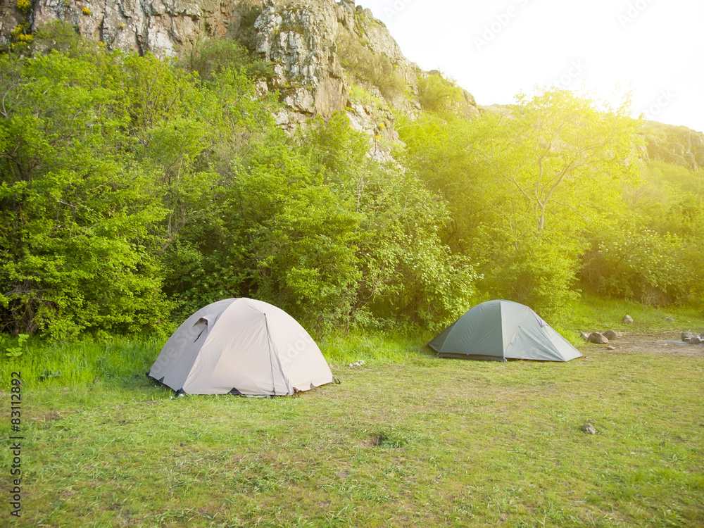 Two tents stand in a forest with mountains in background.