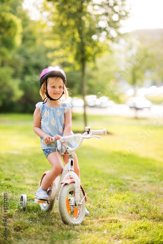 Fototapeta Naklejka Na Ścianę i Meble -  Girl in pink helmet sitting on her bike in a sunny city park