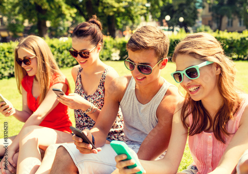 smiling friends with smartphones sitting on grass