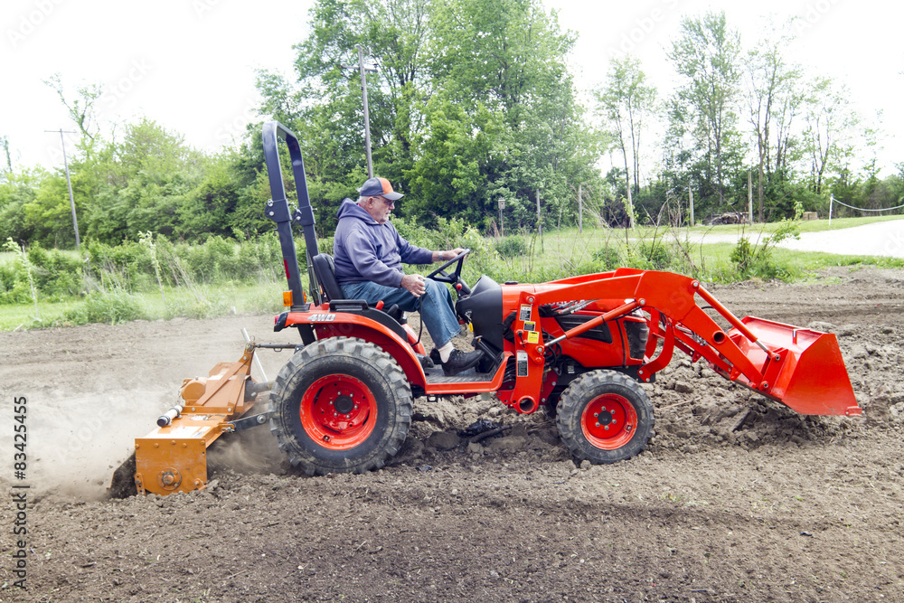 Older Gentleman Tilling His Garden With A Compact Tractor Older Gentleman Tilling His Garden With A Compact Tractor