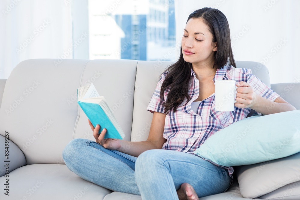 Obraz premium Pretty brunette reading and holding a cup of tea on couch