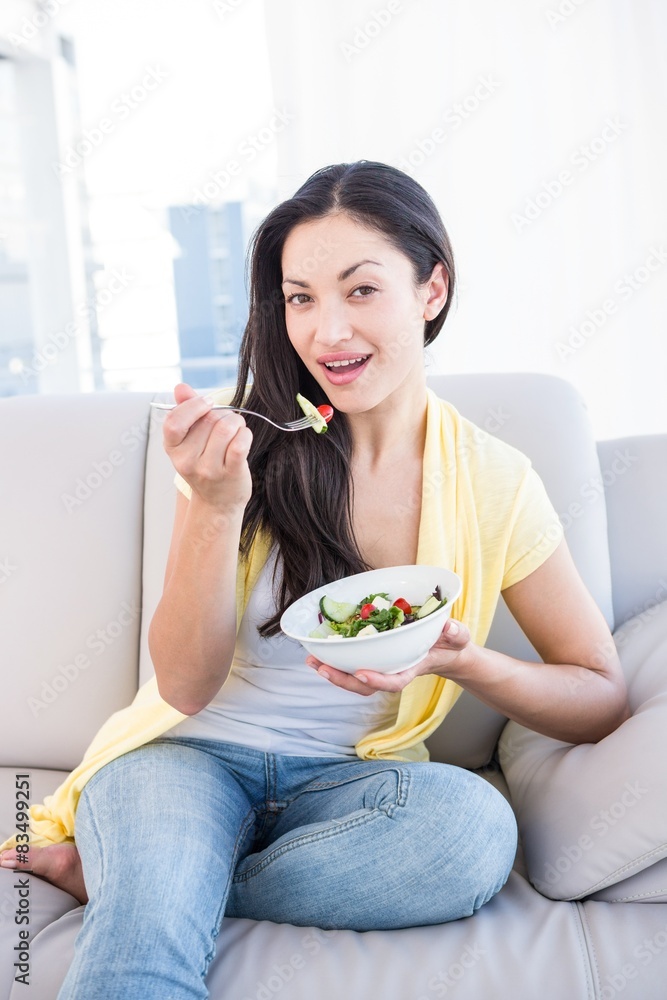 Pretty brunette looking at camera and eating salad on couch