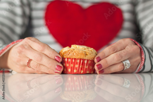 Close-up of a Woman wearing a jumper with a big red heart sitting at a table holding a muffin