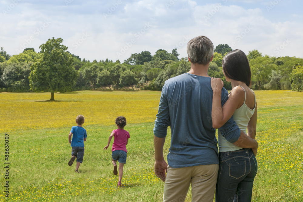 back view of a couple holding each other and contemplating a landscape while their two children running in the grass back view of a couple holding each other and contemplating a landscape while their two children running in the grass