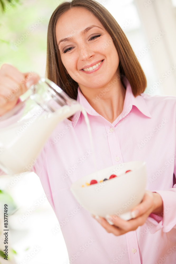 Smiling woman is pouring a milk in corn flakes