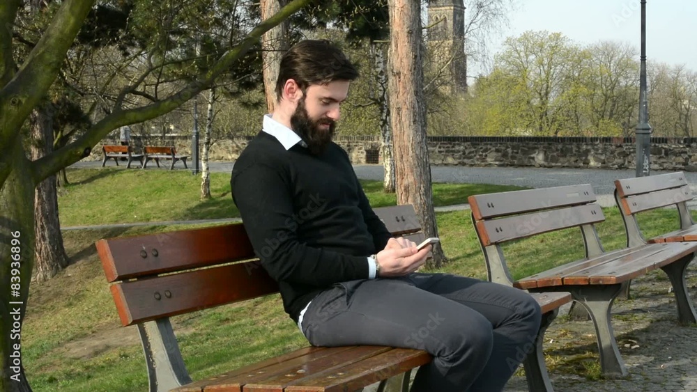 young handsome man with full-beard (hipster) sits on bench and works on mobile phone - park (nature) young handsome man with full-beard (hipster) sits on bench and works on mobile phone - park (nature)