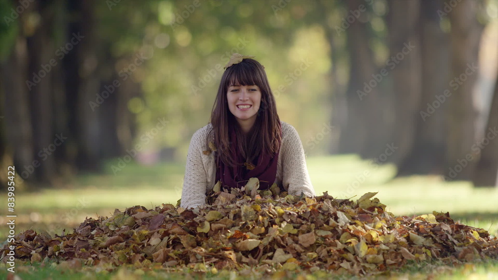 A grown woman sitting in a pile of leaves in the park smiles and throws them up into the air, in slow motion A grown woman sitting in a pile of leaves in the park smiles and throws them up into the air, in slow motion