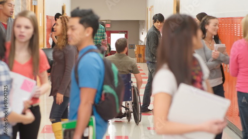 A teacher in a wheelchair talks a groups of students standing in the hallway