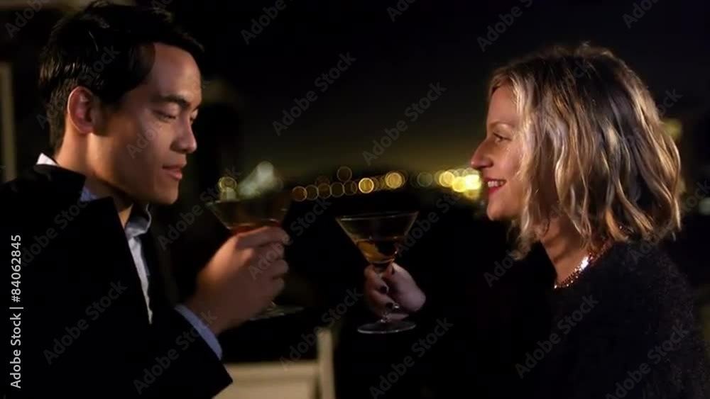 A man and woman stand and talk with each other on a rooftop at night, they then toast and drink A man and woman stand and talk with each other on a rooftop at night, they then toast and drink