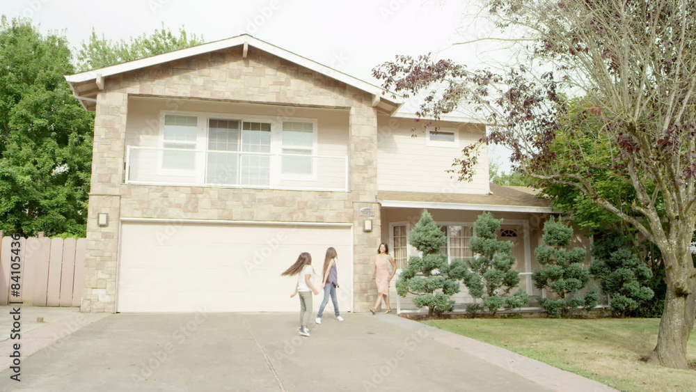 Two young sisters and their mother walk out of the front door of their house and walk down the driveway to smile and wave at the camera