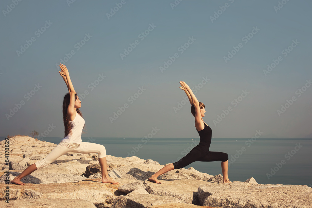 Beautiful young women practicing yoga Virabhadrasana pose on sea beach on the rocks outdoor Beautiful young women practicing yoga Virabhadrasana pose on sea beach on the rocks outdoor