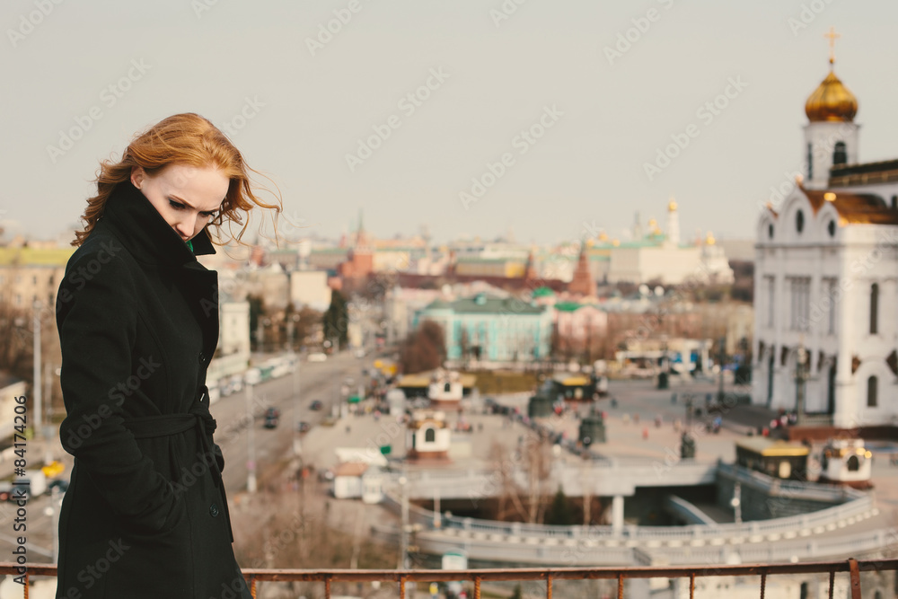Beautiful young woman in a black coat with red hair near the church in Moscow, capital of Russia