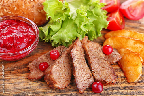 Beef with cranberry sauce, roasted potato slices and bun on wooden board background
