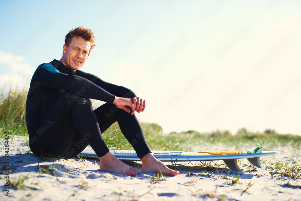 Male Surfer Sits Beside his Surfboard on the Sand