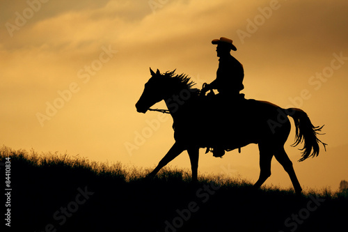 A silhouette of a cowboy and horse walking up a meadow with an orange and yellow background sky.
