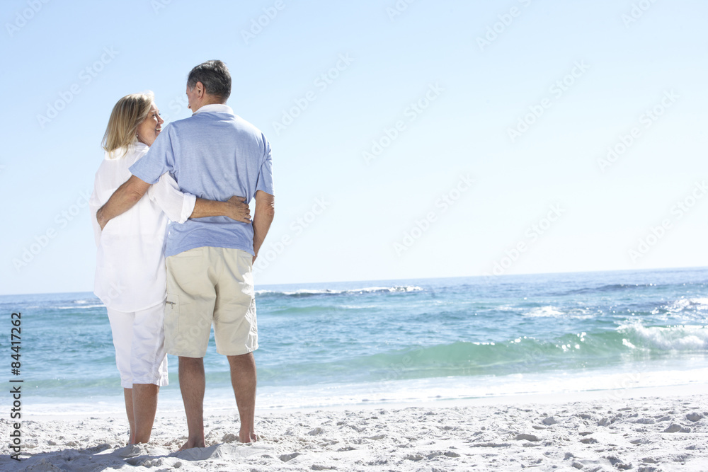 Senior Couple On Holiday Walking Along Sandy Beach Looking Out To Sea
