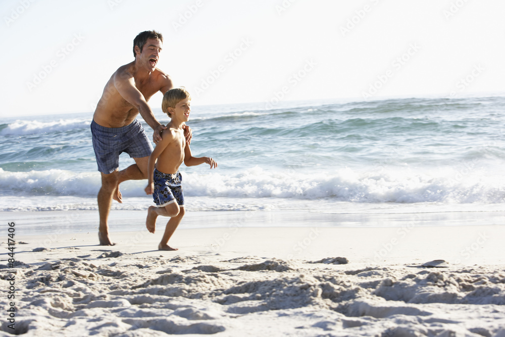 Father And Son Running Along Beach Together Wearing Swimming Costumes Father And Son Running Along Beach Together Wearing Swimming Costumes