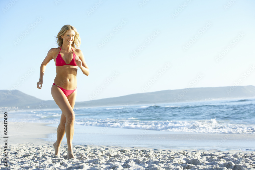 Naklejka premium Young Woman Running Along Sandy Beach On Holiday Wearing Bikini