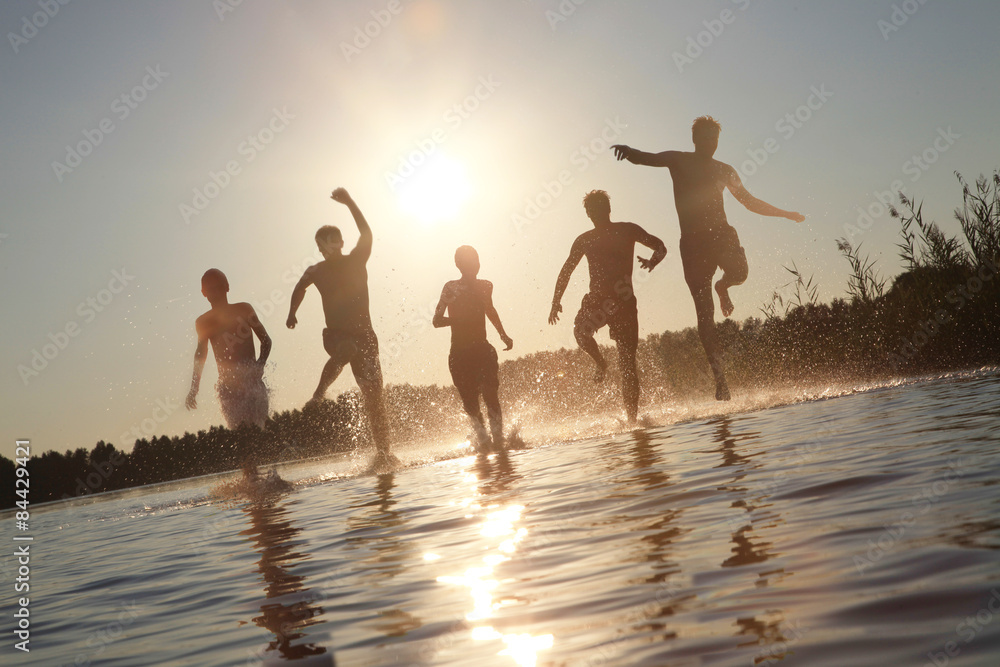Fototapeta premium Glückliche junge Menschen laufen und springen am See beim Sonnenuntergang
