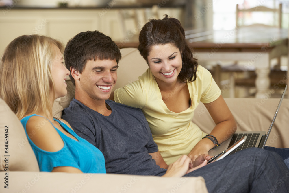 Three Teenagers Sitting On Sofa At Home Using Mobile Phone, Tablet Computer And Laptop Three Teenagers Sitting On Sofa At Home Using Mobile Phone, Tablet Computer And Laptop