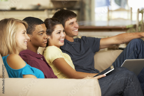 Four Teenagers  Sitting On Sofa At Home Using Tablet Computer And Laptop Whilst Watching TV