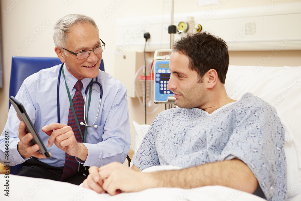 Obraz premium Doctor Sitting By Male Patient's Bed Using Digital Tablet