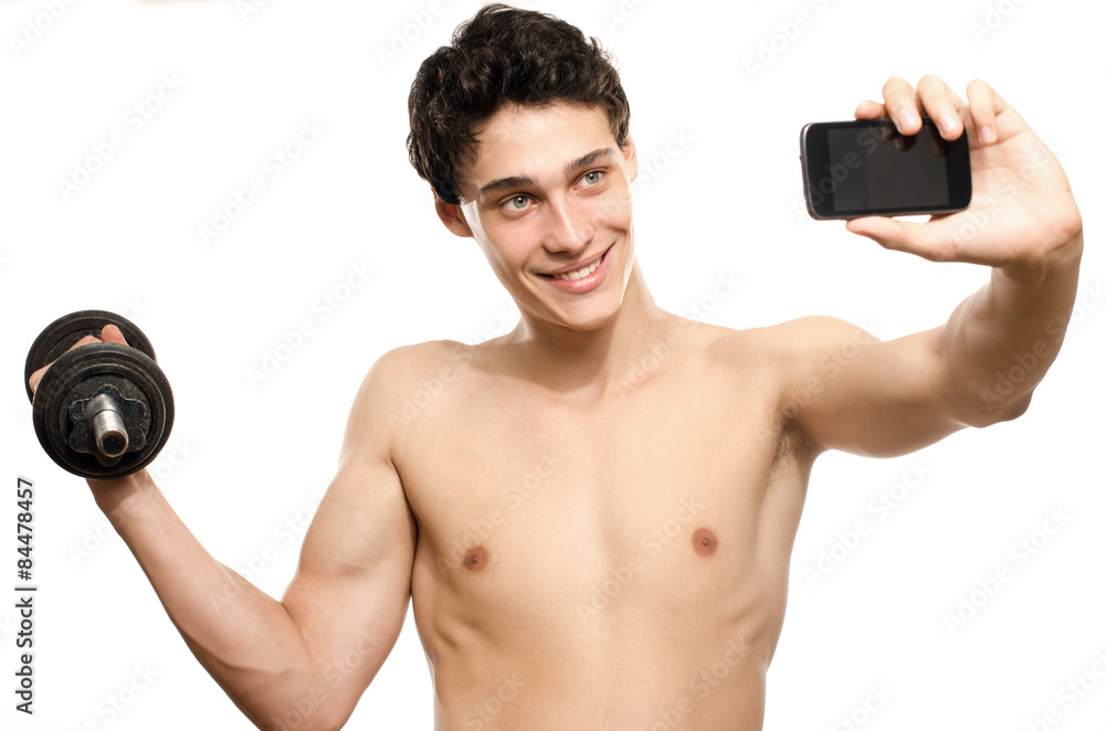 Skinny man taking a selfie with his phone while training his bicep muscle.  Beautiful teenager lifting a dumbbell and taking a photo for  facebook.Anorexic young man training to become stronger Photos