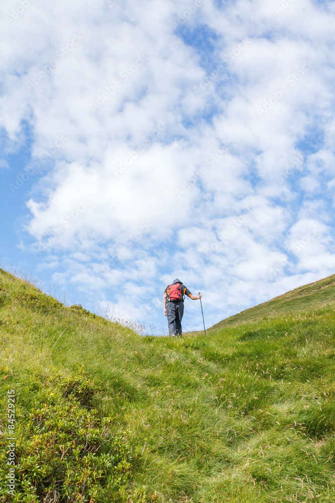 senda verde / Hombre de espaldas subiendo por un sendero de montaña, cielo azul con nubes blancas