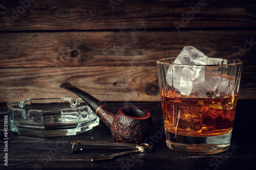 Fotografie Glass of whiskey with ice and pipe on a wooden background.