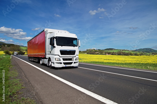 Obraz na płótnie White trucks driving on asphalt road between yellow flowering rapeseed field w salonie