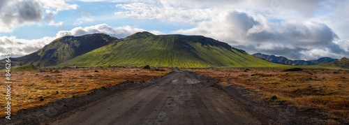 Panoramic view of dirt road leading to green, moss covered volcano mountain under a blue sky with white clouds, Landmannalaugar, Iceland.